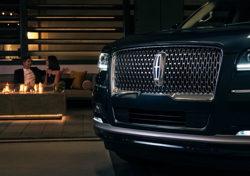 The signature Lincoln grille on the 2022 Lincoln Navigator SUV commands attention as a couple relaxes by a fire beyond | Klaben Lincoln in Kent OH