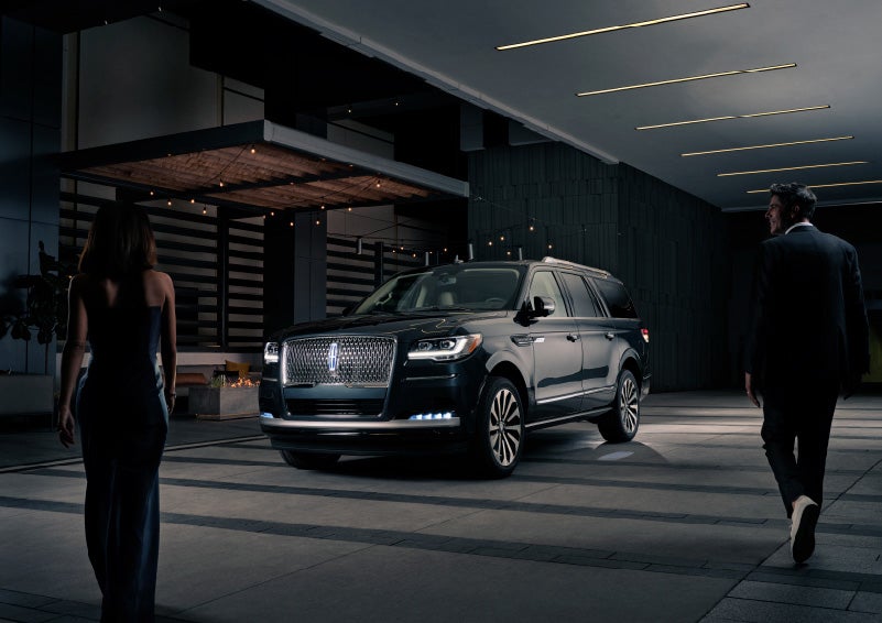 A man and woman in formal wear are approaching a 2022 Lincoln Navigator SUV at night as it lights with the Lincoln Embrace | Klaben Lincoln in Kent OH