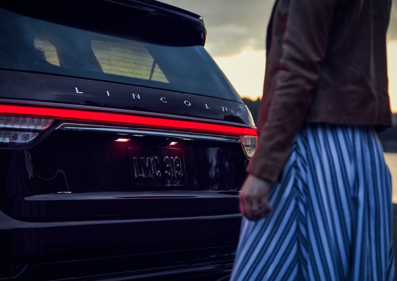 A person is shown near the rear of a 2024 Lincoln Aviator® SUV as the Lincoln Embrace illuminates the rear lights | Klaben Lincoln in Kent OH