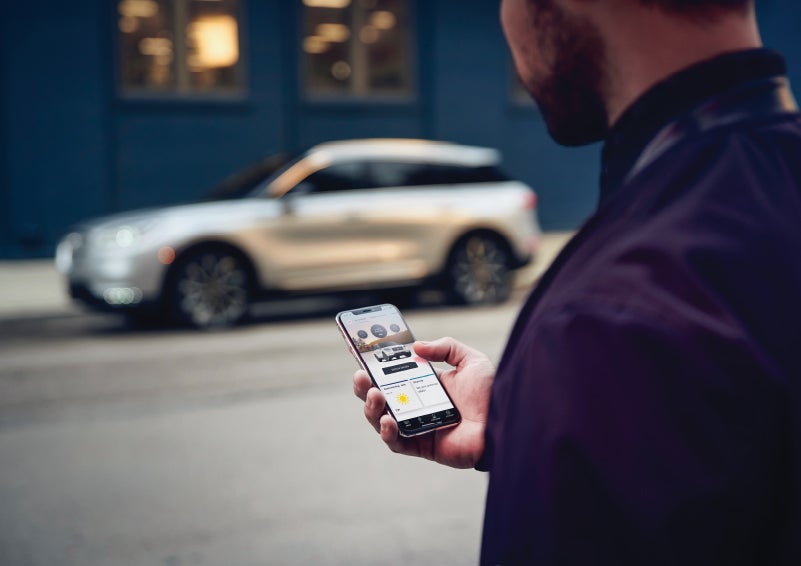 A man is holding his phone looking at the Lincoln Way™ App and we see his 2022 Lincoln Corsair SUV in the background against a blue brick wall | Klaben Lincoln in Kent OH