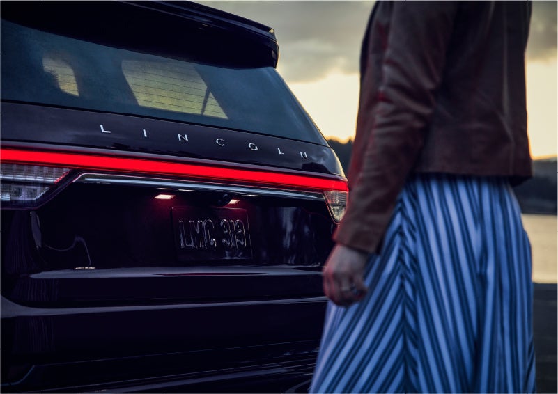 A person is shown near the rear of a 2023 Lincoln Aviator® SUV as the Lincoln Embrace illuminates the rear lights | Klaben Lincoln in Kent OH