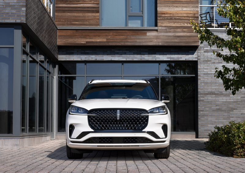 A white 2025 Lincoln Aviator® SUV is shown parked outside a modern building with gray brick and wood accents. | Klaben Lincoln in Kent OH