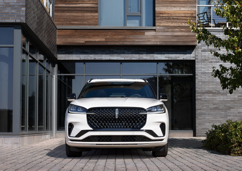 A white 2025 Lincoln Aviator® SUV is shown parked outside a modern building with gray brick and wood accents. | Klaben Lincoln in Kent OH