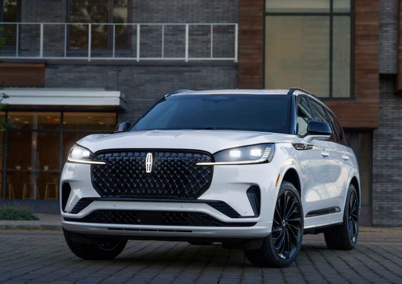 A white 2025 Lincoln Aviator® SUV is shown parked outside a modern building with gray brick and wood accents. | Klaben Lincoln in Kent OH